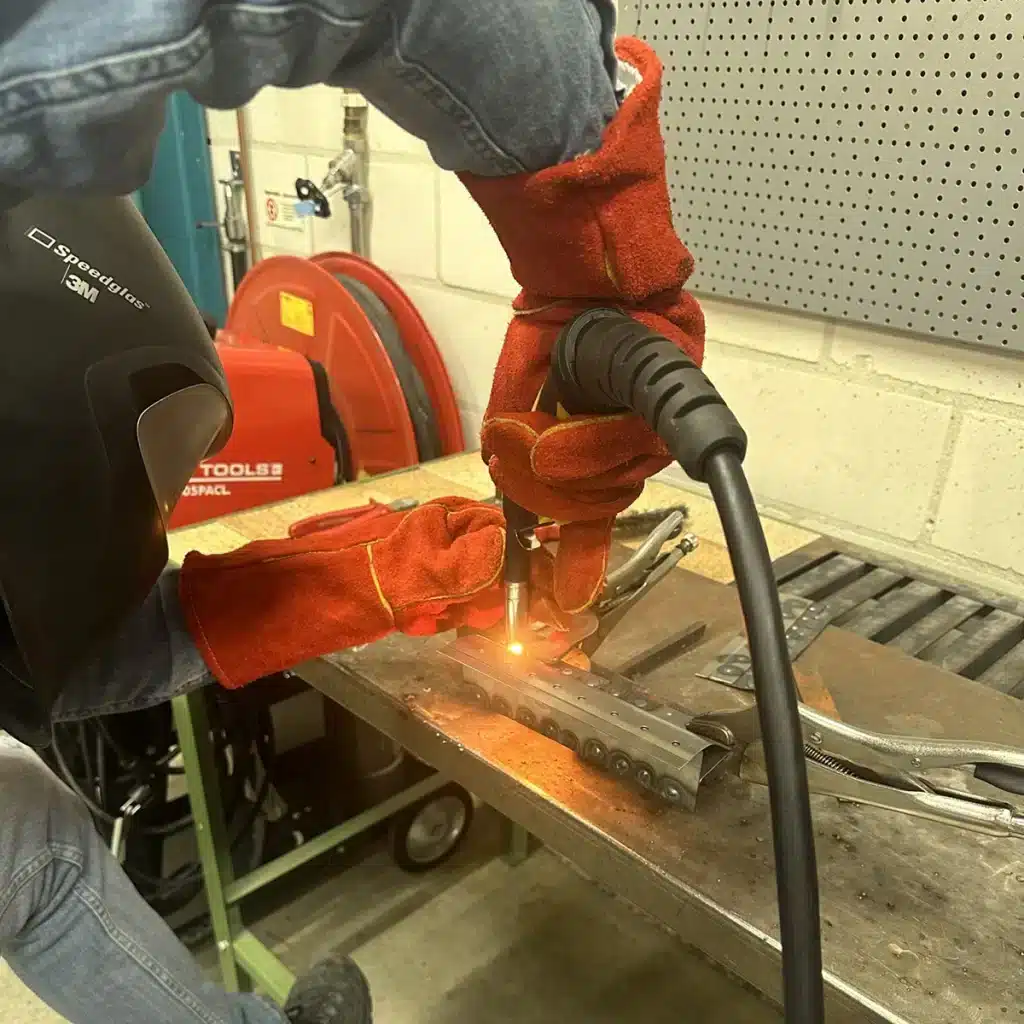 Close-up van een dunne metalen autoplaat die aan een kokerbalk wordt gelast, met zichtbaar lasproces en metalen oppervlakken